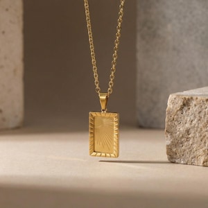 Puede incluir: Un collar con colgante dorado con un colgante rectangular con un diseño de rayos de sol. El colgante cuelga de una delicada cadena. El collar se muestra sobre un fondo neutro.