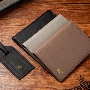 May include: An overhead shot shows a black luggage tag and three wallets in black, gray, and brown. The tag and wallets have gold initials. The items are on a wooden surface.