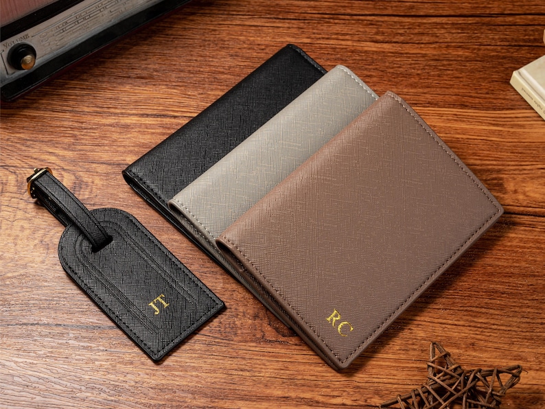 May include: An overhead shot shows a black luggage tag and three wallets in black, gray, and brown. The tag and wallets have gold initials. The items are on a wooden surface.
