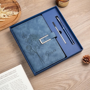May include: A blue notebook with a floral line art design and the name Grace embossed on the cover, featuring a gold-toned metal clasp. The notebook is presented in a matching blue gift box alongside a blue ballpoint pen and a metal pen refill.