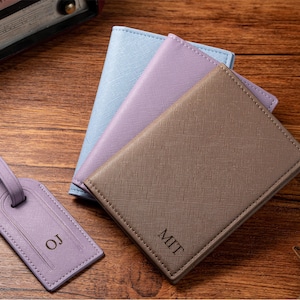 May include: A collection of personalized travel accessories, including a brown passport holder with "MIT" engraved, a lavender luggage tag with "OJ", and two additional passport holders in light blue and lavender.