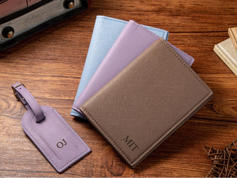May include: A collection of personalized travel accessories, including a brown passport holder with "MIT" engraved, a lavender luggage tag with "OJ", and two additional passport holders in light blue and lavender.
