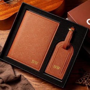May include: A brown leather passport holder and luggage tag set in a black gift box. The set includes a passport holder and a luggage tag, both with the initials "BW" in gold. The set is presented on a wooden surface.