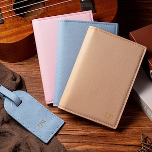 May include: A collection of travel accessories, including a light blue luggage tag with gold initials, and three passport holders in pink, light blue, and tan. A ukulele and books are in the background. The items are on a wooden surface.