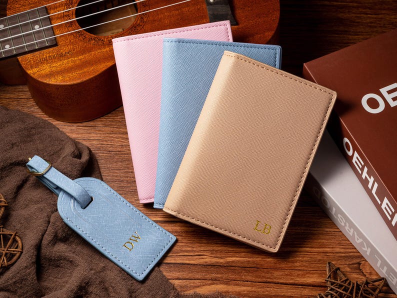 May include: A collection of travel accessories, including a light blue luggage tag with gold initials, and three passport holders in pink, light blue, and tan. A ukulele and books are in the background. The items are on a wooden surface.