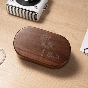 K&ouml;nnte beinhalten: Eine dunkelbraune, ovale Holzbox mit floraler Gravur und dem Namen "Layla" auf dem Deckel. Ein wei&szlig;es Perlenarmband, ein Silberring und ein wei&szlig;er Retro-Plattenspieler sind ebenfalls zu sehen.