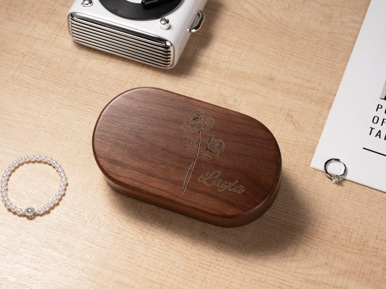 K&ouml;nnte beinhalten: Eine dunkelbraune, ovale Holzbox mit floraler Gravur und dem Namen "Layla" auf dem Deckel. Ein wei&szlig;es Perlenarmband, ein Silberring und ein wei&szlig;er Retro-Plattenspieler sind ebenfalls zu sehen.