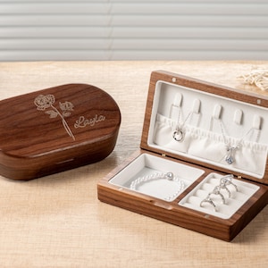 K&ouml;nnte beinhalten: Eine offene Holzschmuckschatulle mit Halsketten und Ringen. Die Schatulle ist braun mit wei&szlig;em Innenraum. Daneben eine geschlossene, ovale Schatulle mit Blumendesign und dem Namen "Layla". Der Schmuck ist silberfarben.