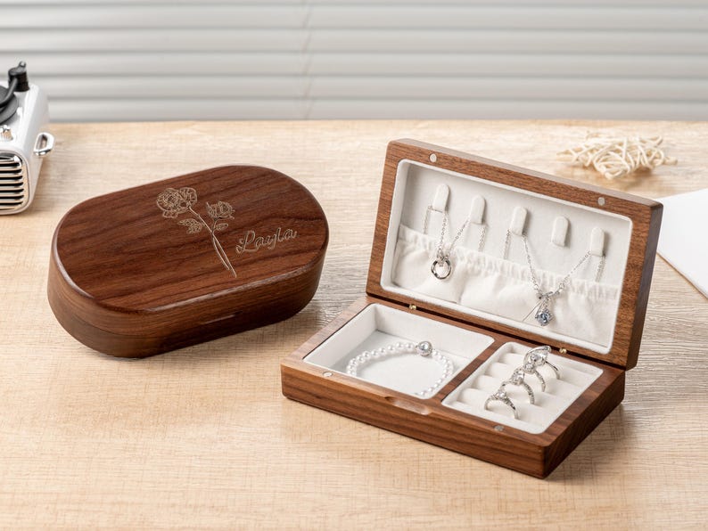 K&ouml;nnte beinhalten: Eine offene Holzschmuckschatulle mit Halsketten und Ringen. Die Schatulle ist braun mit wei&szlig;em Innenraum. Daneben eine geschlossene, ovale Schatulle mit Blumendesign und dem Namen "Layla". Der Schmuck ist silberfarben.