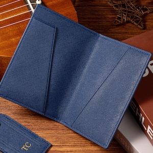 May include: A navy blue leather passport holder is open, revealing interior pockets. A matching luggage tag with gold initials lies nearby. The background includes a ukulele and a book.