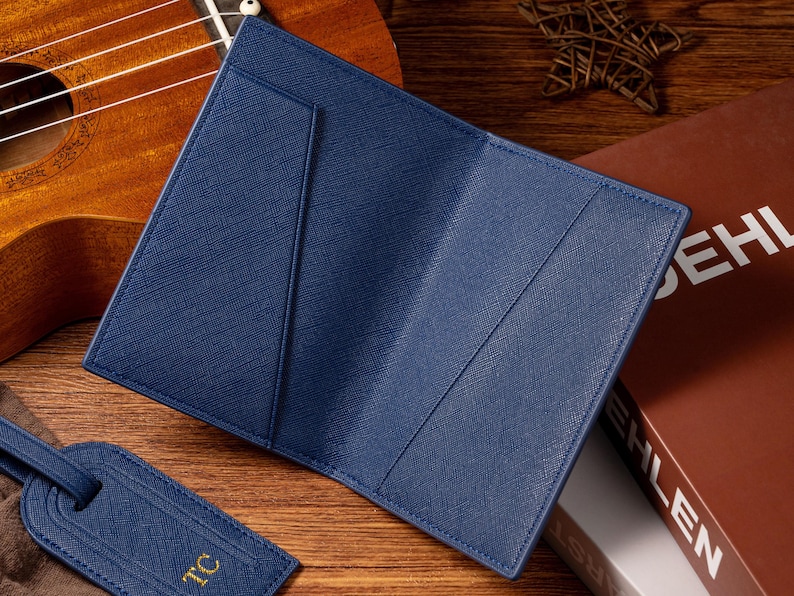 May include: A navy blue leather passport holder is open, revealing interior pockets. A matching luggage tag with gold initials lies nearby. The background includes a ukulele and a book.