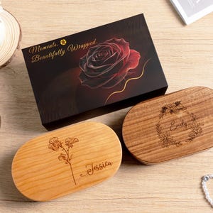 May include: A black rectangular gift box featuring a red rose graphic and the text Moments, Beautifully Wrapped, alongside two oval wooden boxes. One light wood box is engraved with a floral stem and the name Jessica, while the darker wood box features a floral wreath and the name Emily.