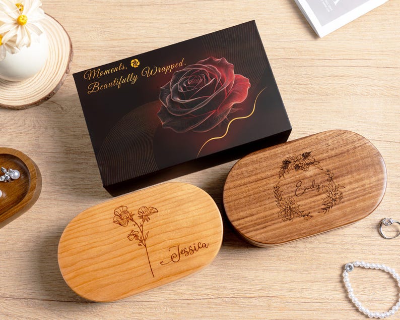May include: A black rectangular gift box featuring a red rose graphic and the text Moments, Beautifully Wrapped, alongside two oval wooden boxes. One light wood box is engraved with a floral stem and the name Jessica, while the darker wood box features a floral wreath and the name Emily.