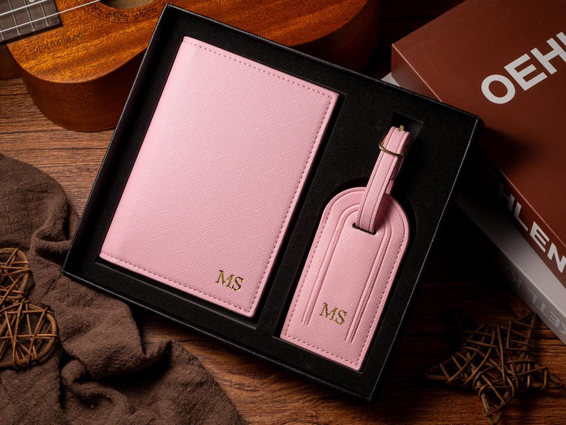 May include: A pink passport holder and luggage tag set, presented in a black gift box. Both items are embossed with the initials "MS" in gold. The set is made of a textured material and includes a luggage tag with a buckle.