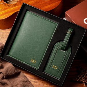 May include: A dark green passport holder and luggage tag set in a black gift box. The passport holder and luggage tag have gold initials. The set is displayed on a wooden surface with a ukulele and a book in the background.