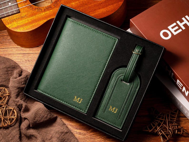 May include: A dark green passport holder and luggage tag set in a black gift box. The passport holder and luggage tag have gold initials. The set is displayed on a wooden surface with a ukulele and a book in the background.