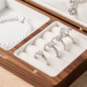 K&ouml;nnte beinhalten: Eine Holzschmuckschatulle mit wei&szlig;em Innenraum, die ein Perlenarmband, drei silberne Ringe mit klaren Edelsteinen und eine silberne Anh&auml;ngerkette enth&auml;lt. Die Schatulle ist ge&ouml;ffnet und zeigt den Schmuck.