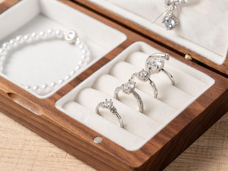 K&ouml;nnte beinhalten: Eine Holzschmuckschatulle mit wei&szlig;em Innenraum, die ein Perlenarmband, drei silberne Ringe mit klaren Edelsteinen und eine silberne Anh&auml;ngerkette enth&auml;lt. Die Schatulle ist ge&ouml;ffnet und zeigt den Schmuck.