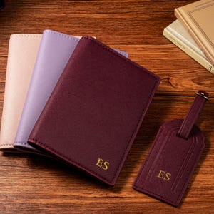 May include: Three passport holders in blush, lavender, and burgundy, with a matching burgundy luggage tag. The burgundy items are embossed with the initials "ES" in gold. The items are arranged on a wooden surface.