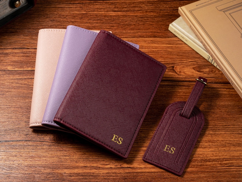 May include: Three passport holders in blush, lavender, and burgundy, with a matching burgundy luggage tag. The burgundy items are embossed with the initials "ES" in gold. The items are arranged on a wooden surface.