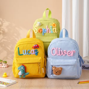 May include: Three corduroy backpacks in yellow, light green, and light blue. The yellow bag features the name Lucas with a red car patch and green dinosaur. The green bag displays Sophia with a bee and flower. The blue bag shows Oliver with a bear.