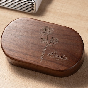 K&ouml;nnte beinhalten: Eine dunkelbraune, ovale Holzbox mit einer Rosen-Gravur und dem Namen "Layla" in Schreibschrift. Die Box steht auf einer hellbraunen Holzoberfl&auml;che. Im Hintergrund befindet sich ein wei&szlig;es und silbernes Objekt.