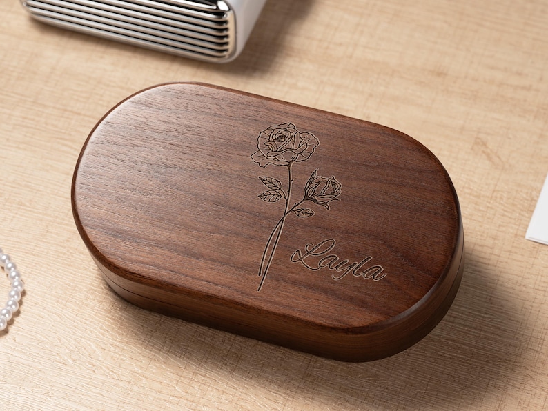 K&ouml;nnte beinhalten: Eine dunkelbraune, ovale Holzbox mit einer Rosen-Gravur und dem Namen "Layla" in Schreibschrift. Die Box steht auf einer hellbraunen Holzoberfl&auml;che. Im Hintergrund befindet sich ein wei&szlig;es und silbernes Objekt.