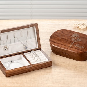 K&ouml;nnte beinhalten: Eine braune Holzschmuckschatulle, ge&ouml;ffnet, um Halsketten, ein Armband und Ringe zu zeigen. Die Schatulle hat einen hellen Innenraum. Daneben eine geschlossene, passende Holzschatulle mit floralem Design und dem Namen "Layla".