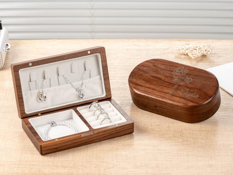 K&ouml;nnte beinhalten: Eine braune Holzschmuckschatulle, ge&ouml;ffnet, um Halsketten, ein Armband und Ringe zu zeigen. Die Schatulle hat einen hellen Innenraum. Daneben eine geschlossene, passende Holzschatulle mit floralem Design und dem Namen "Layla".