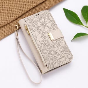 May include: Cream-colored phone wallet with a floral design. The wallet features a zipper, wrist strap, and a gold-colored clasp. The floral pattern is etched into the surface, creating a textured look. The wallet is made of a soft material.