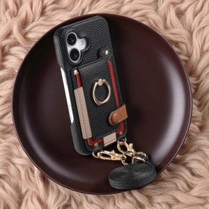 May include: Black leather phone case with a cardholder, ring holder, and detachable black strap. The case features a gold ring, red and beige accents, and a small brown leather detail. The phone case is on a dark brown plate.