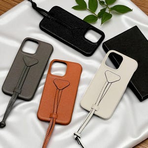 以下が含まれることがあります： 黒、グレー、オレンジ、オフホワイトのレザー製携帯電話ケース。各ケースには、三角形のアクセントとリストループが付いたストラップが付いています。ケースは白い表面に表示されています。測定値は適用されません。