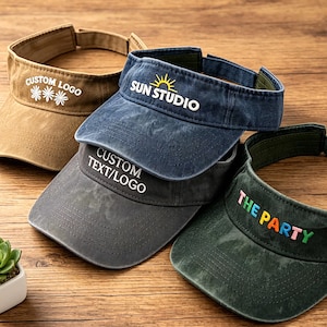 Pode incluir: Quatro viseiras em várias cores: bege, azul marinho, cinza e verde escuro. Cada viseira apresenta textos ou logotipos personalizados, incluindo "CUSTOM LOGO", "SUN STUDIO", "CUSTOM TEXT/LOGO" e "THE PARTY". As viseiras estão dispostas sobre uma superfície de madeira.