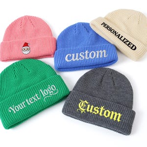 Op de afbeelding: Vijf geribbelde gebreide mutsen in verschillende kleuren: roze met een kerstmanontwerp, blauw met "custom" borduursel, beige met "PERSONALIZED" tekst, groen met "Your text/logo" en grijs met "Custom" borduursel.
