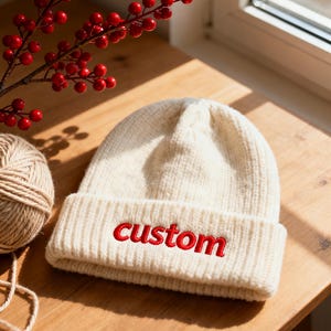 May include: A cream-coloured knitted beanie hat with the word "custom" embroidered in red. The hat is folded at the brim and rests on a wooden surface. A ball of twine and a branch with red berries are also visible.