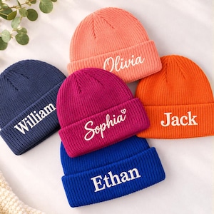 Op de afbeelding: Vijf gebreide mutsen in koraal, marineblauw, magenta, oranje en koningsblauw. Elke muts heeft een omgeslagen boord met een wit geborduurde naam: Olivia, William, Sophia met een hartje, Jack en Ethan in verschillende lettertypes.
