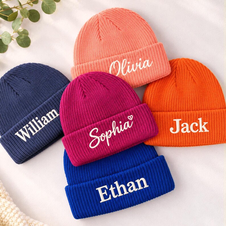Op de afbeelding: Vijf gebreide mutsen in koraal, marineblauw, magenta, oranje en koningsblauw. Elke muts heeft een omgeslagen boord met een wit geborduurde naam: Olivia, William, Sophia met een hartje, Jack en Ethan in verschillende lettertypes.