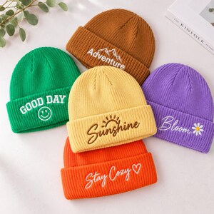 Op de afbeelding: Een collectie van vijf gebreide mutsen in groen, bruin, geel, oranje en paars. De groene muts toont GOOD DAY met een smiley, bruin toont Adventure met bergen, geel toont Sunshine met een zon, paars toont Bloom met een madeliefje en oranje toont Stay Cozy met een hartje.