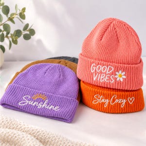 Op de afbeelding: Een collectie van vier geribbelde gebreide mutsen in koraal, oranje, paars en bruin. De koraalkleurige muts heeft wit geborduurd GOOD VIBES-tekst met een madeliefje. De oranje muts heeft witte tekst met Stay Cozy en een hartje.