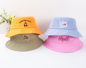 Op de afbeelding: Vier kleurrijke bucket hats. Eén is oranje met "CAMPING CLUB" en een kampvuurafbeelding. Een andere is lichtblauw met "I Love Dad". Een derde is roze met "Bride". De laatste is beige met "love myself".