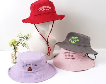 Op de afbeelding: Een verzameling vissershoeden in verschillende kleuren, waaronder rood, grijs en lavendel. De rode hoed is geborduurd met "CUSTOM EST. 2025". De grijze hoed heeft een palmbomenontwerp en de lavendelkleurige hoed toont "CAMPING CLUB".