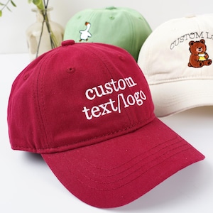 May include: A close-up of three baseball caps. The frontmost cap is burgundy with the words "custom text/logo" embroidered in white. A light green cap with a white goose graphic and a cream cap with a bear graphic are behind it.