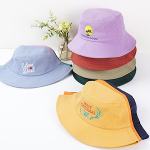 May include: A selection of bucket hats in shades of blue, purple, and yellow. Some hats have embroidered text like "I Love Dad" and "Merry Christmas", and a custom logo. These casual hats are suitable for outdoor activities and come in a range of colours.
