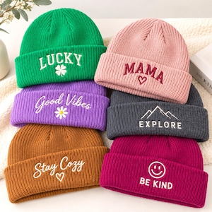 Op de afbeelding: Een collectie van zes geribbelde gebreide mutsen in groen, roze, paars, bruin, donkergrijs en magenta. Elke muts is voorzien van geborduurde tekst en iconen: LUCKY met een klavertje, MAMA met een hartje, Good Vibes met een madeliefje, EXPLORE met bergen, Stay Cozy met een hartje en BE KIND met een smiley.