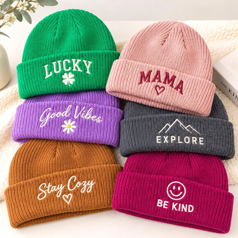 Op de afbeelding: Een collectie van zes geribbelde gebreide mutsen in groen, roze, paars, bruin, donkergrijs en magenta. Elke muts is voorzien van geborduurde tekst en iconen: LUCKY met een klavertje, MAMA met een hartje, Good Vibes met een madeliefje, EXPLORE met bergen, Stay Cozy met een hartje en BE KIND met een smiley.