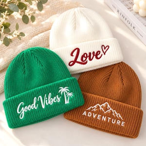 Op de afbeelding: Drie gebreide mutsen in wit, groen en bruin. De witte muts heeft de rode geborduurde tekst "Love" met een hartje. De groene muts heeft de witte geborduurde tekst "Good Vibes" en palmbomen. De bruine muts heeft de witte geborduurde tekst "ADVENTURE" en een bergafbeelding.