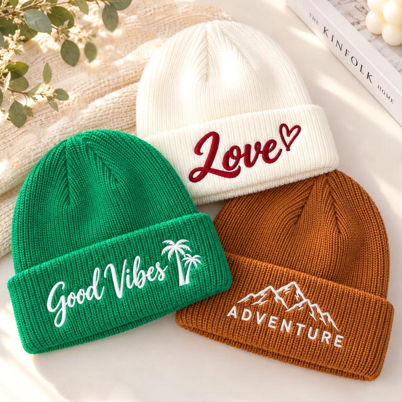 Op de afbeelding: Drie gebreide mutsen in wit, groen en bruin. De witte muts heeft de rode geborduurde tekst "Love" met een hartje. De groene muts heeft de witte geborduurde tekst "Good Vibes" en palmbomen. De bruine muts heeft de witte geborduurde tekst "ADVENTURE" en een bergafbeelding.