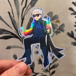 Op de afbeelding: Holografische sticker met een stripfiguur met wit haar, een zonnebril en een zwarte jas, zittend in een stoel. Het personage houdt een zwaard en een blikje vast, met regenboogaccenten. De sticker wordt door een hand vastgehouden.