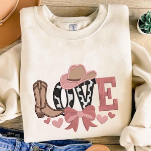 Könnte beinhalten: Cremefarbenes Sweatshirt mit einem gestickten Western-Design. Das Design umfasst einen Cowboyhut, einen Cowboy-Stiefel, eine Schleife, Herzen und das Wort "LOVE" in Rosa und Schwarz.