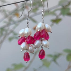 Könnte beinhalten: Ohrringe zum Hängen mit Anhängern aus weißen Perlen und tropfenförmigen, pinkfarbenen Perlen. Die Ohrringe haben silberfarbene Haken und Ketten, die einen Kaskadeneffekt erzeugen. Der Hintergrund ist unscharf.
